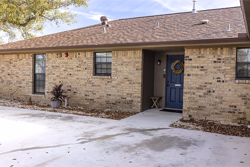 Brick senior living facility exterior with blue front door, wreath, and potted plants near entrance.