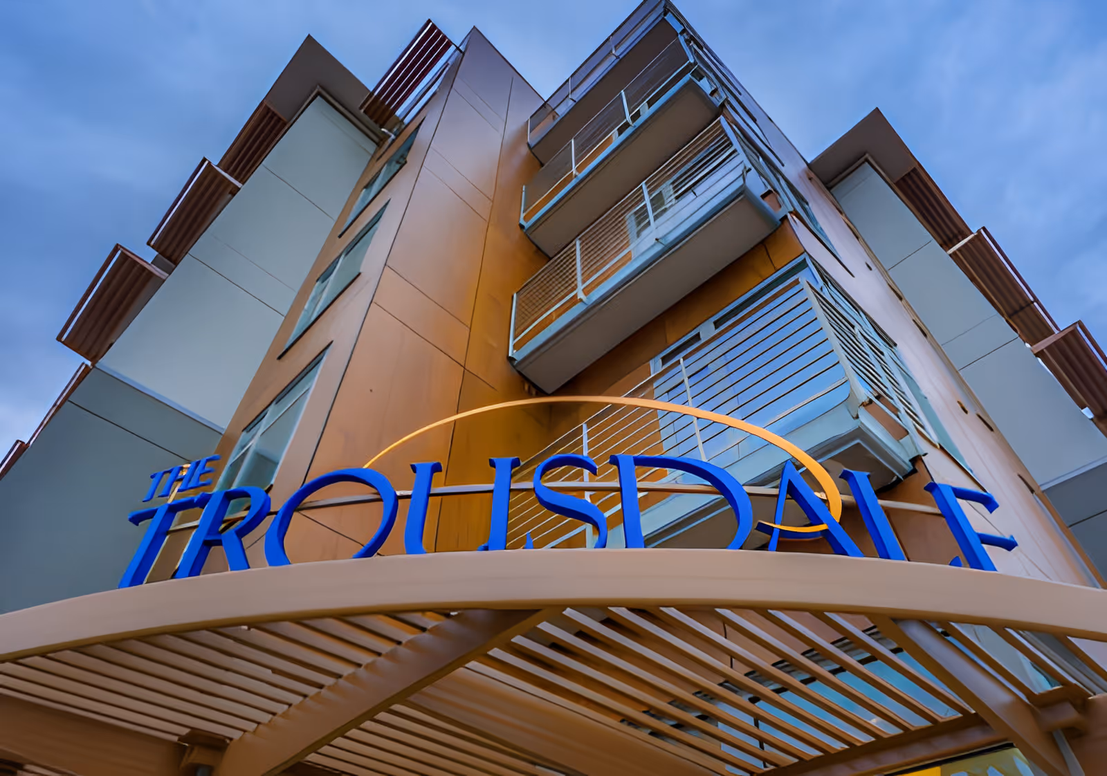 Upward view of the exterior corner of a modern multi-story building with balconies and a sign that reads 'The Trousdale' in blue letters above an entrance canopy.