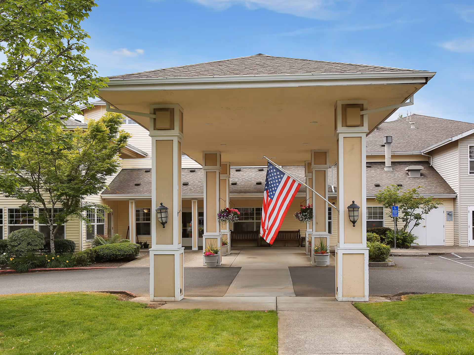 Entrance of Prestige Senior Living Huntington Terrace featuring a covered drop-off area with four beige columns, an American flag hanging in the center, potted plants, and a well-maintained lawn and trees surrounding the building.
