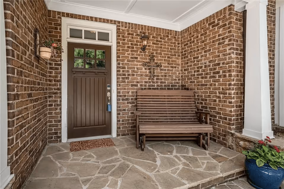 Front porch with a brown wooden door set in brick walls, a wooden bench, and a blue potted plant.