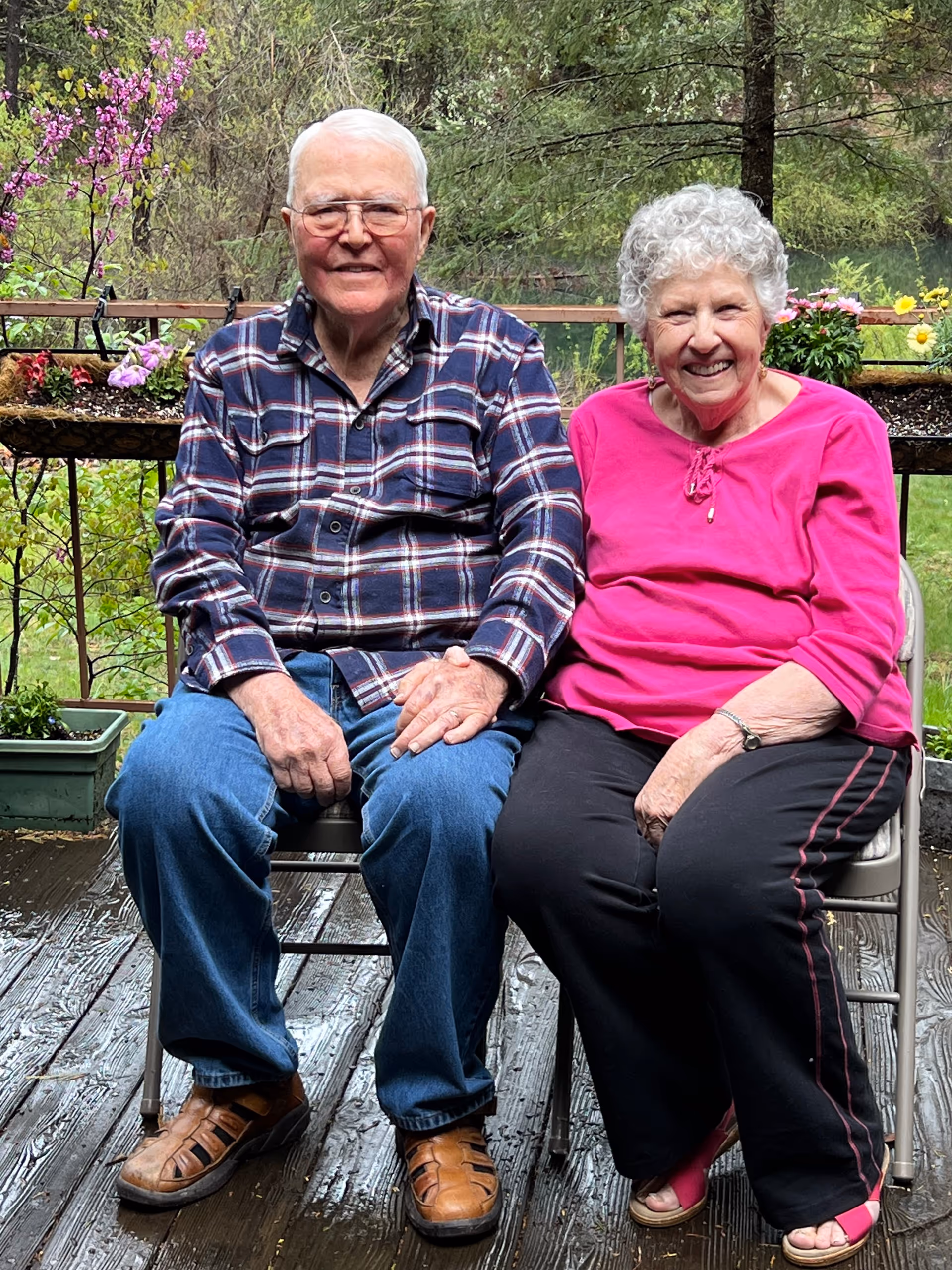 Two elderly individuals sit side-by-side on a wooden outdoor deck with potted flowers.