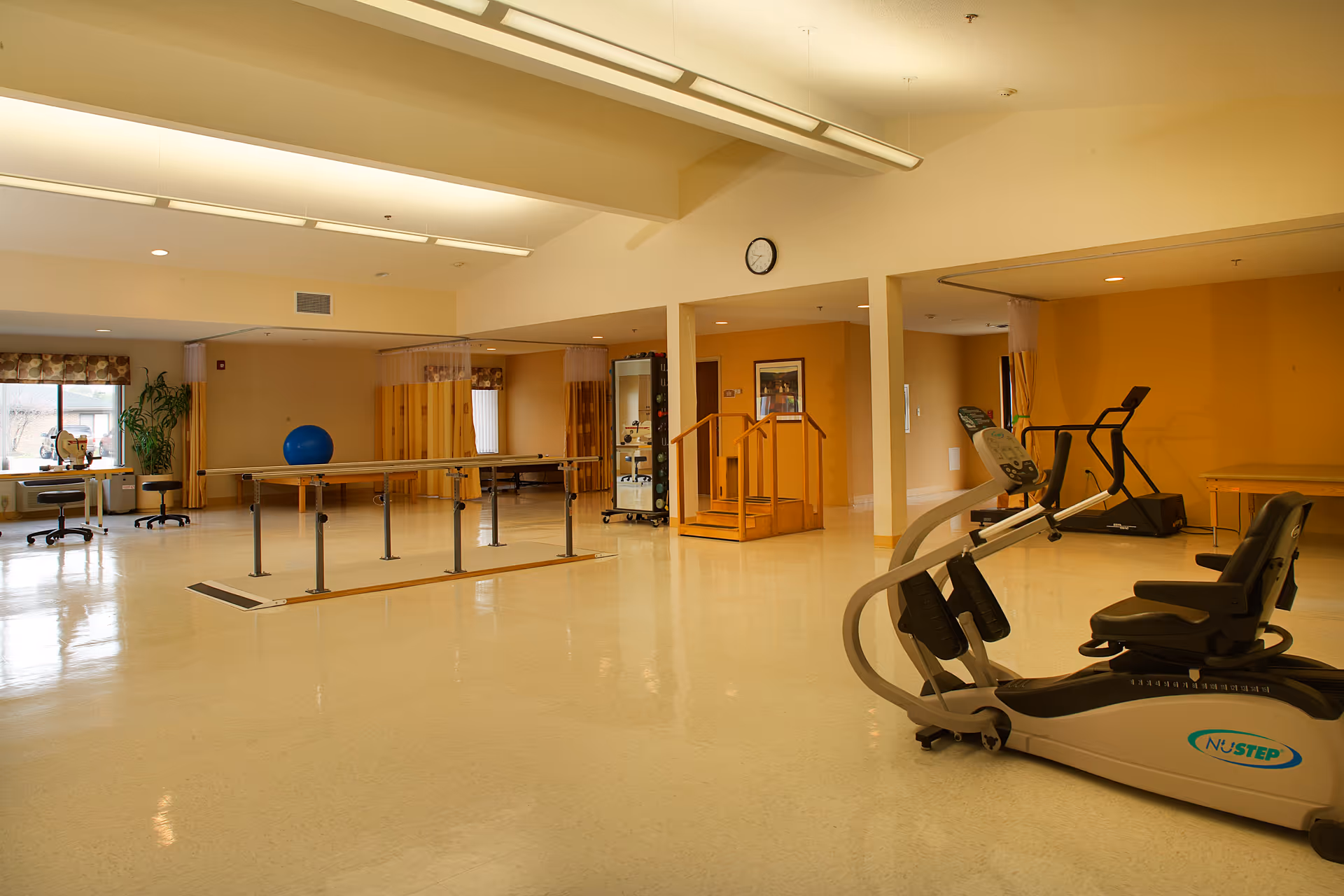 A spacious rehabilitation or physical therapy room with exercise equipment including a recumbent bike and treadmill, parallel bars for walking practice, a small set of stairs with handrails, and a large blue exercise ball. The room has beige walls, a clock on the wall, and large windows letting in natural light.