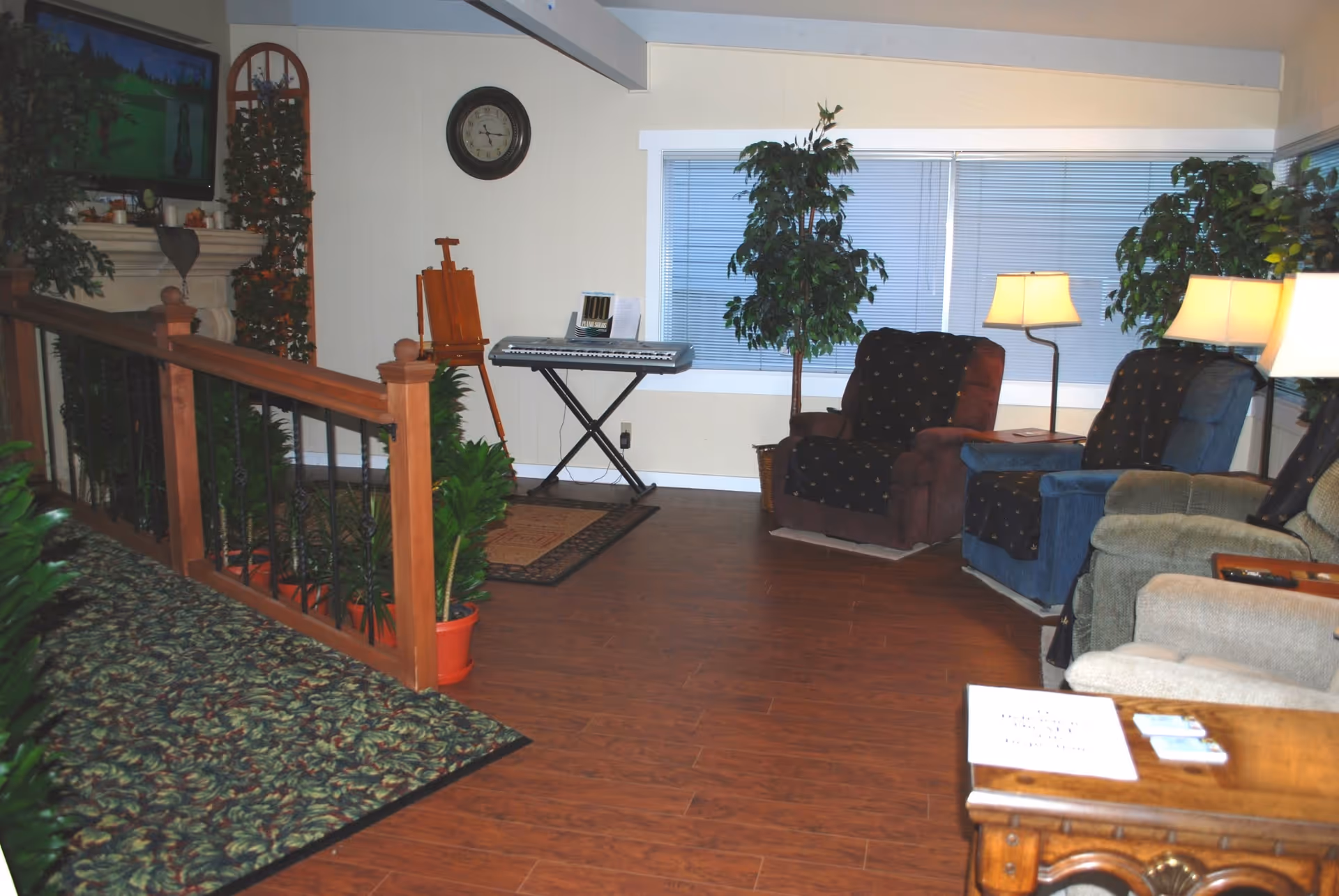 A cozy senior living room with seating, plants, a keyboard on a stand, and staircase railing.
