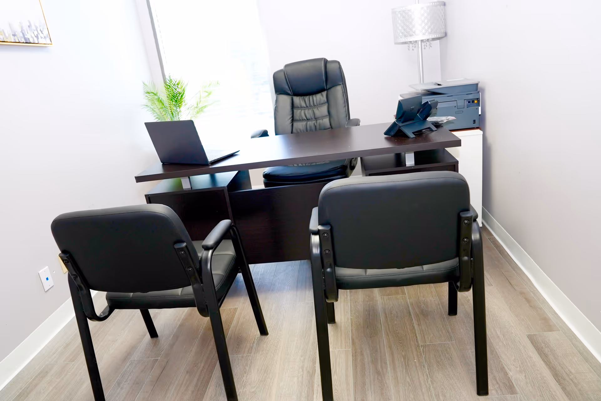 Office setup with desk, laptop, chairs, printer, and a plant in neutral room.