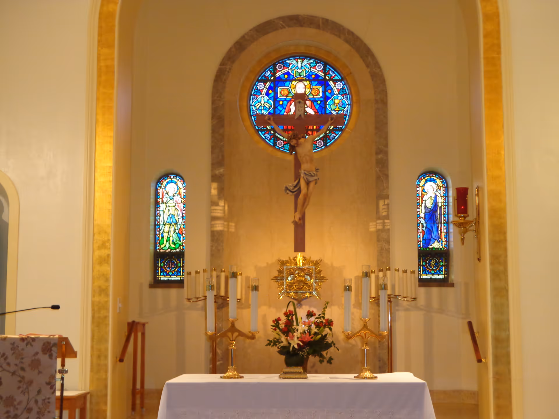 Interior chapel with altar, crucifix, floral arrangement, candlesticks, and colorful stained glass windows.
