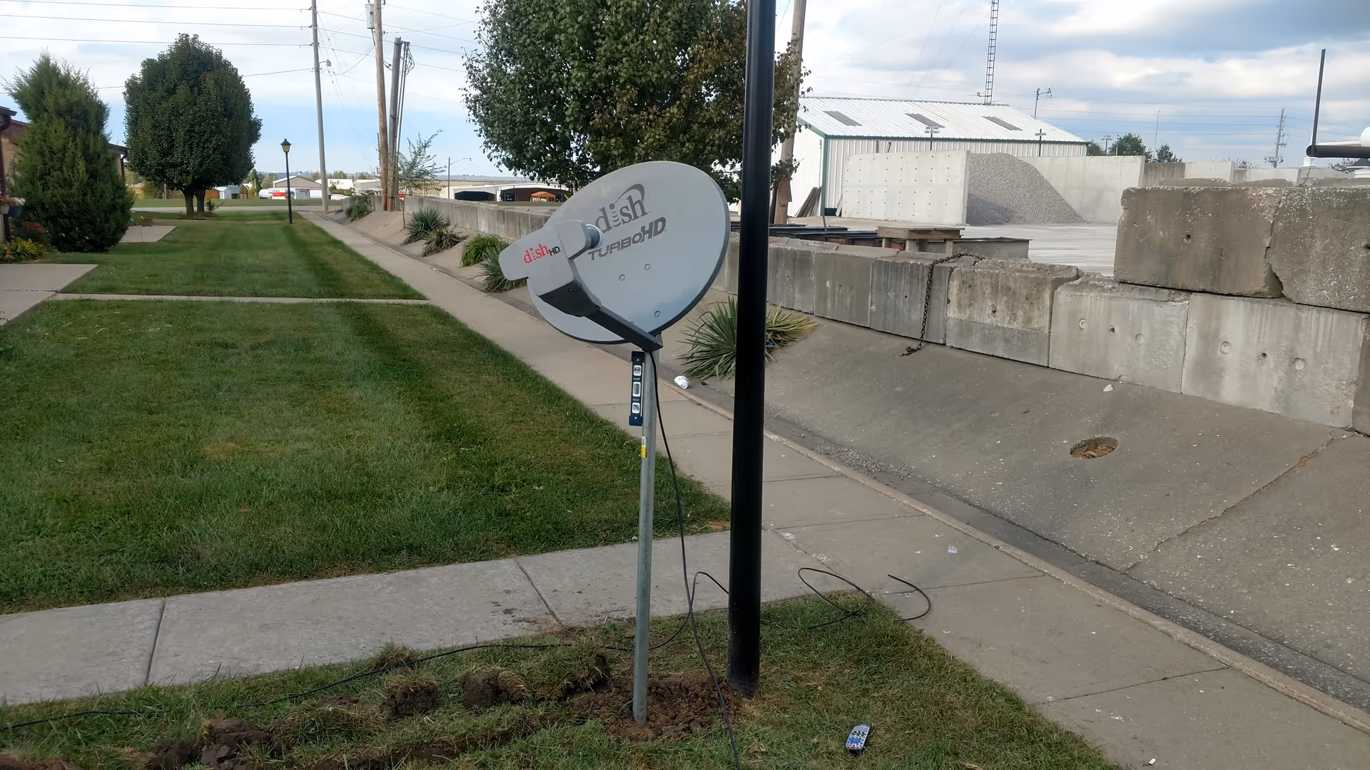 Outdoor sidewalk and grassy strip with a satellite dish mounted beside a concrete barrier.