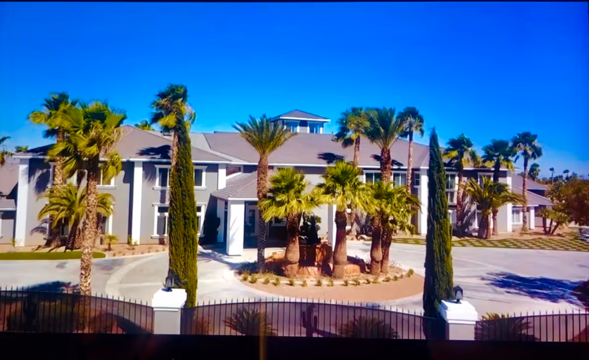Two-story senior living facility building with palm trees and a gated circular driveway under a clear blue sky.