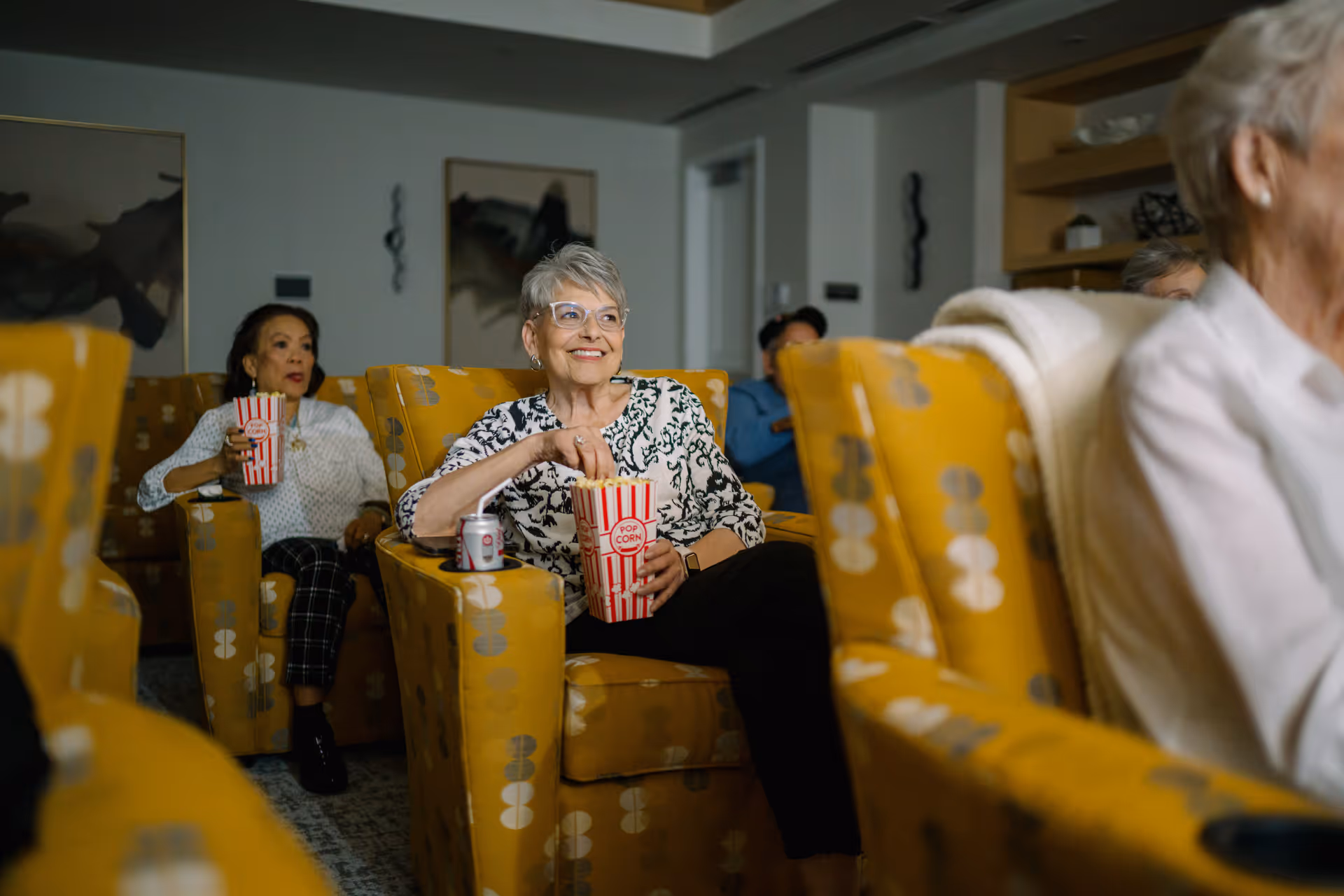 Several elderly women sitting in yellow patterned armchairs in a cozy room, enjoying popcorn and soda while watching something, likely a movie or presentation.