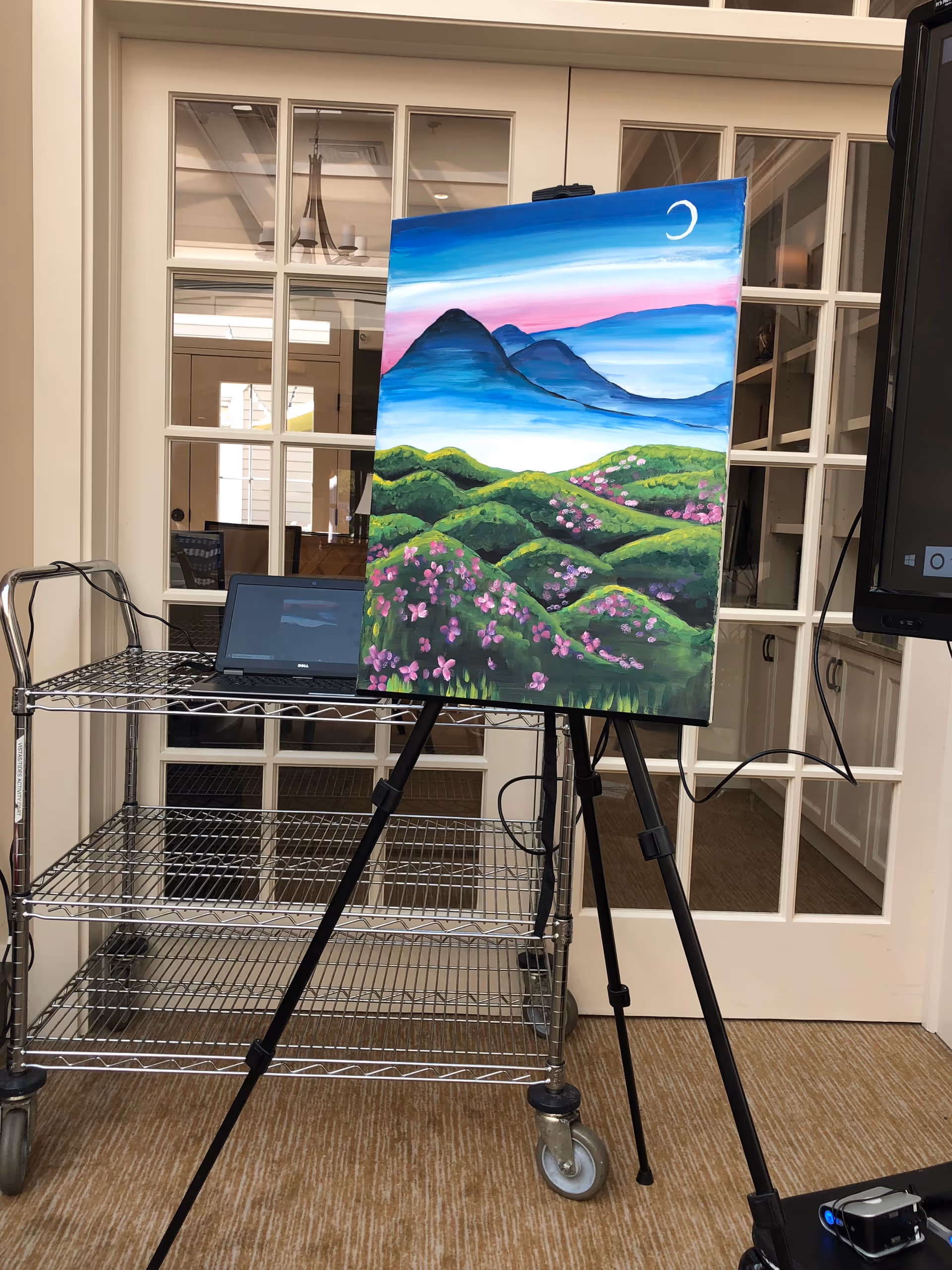 Colorful landscape painting on an easel inside a room beside a metal rolling cart.