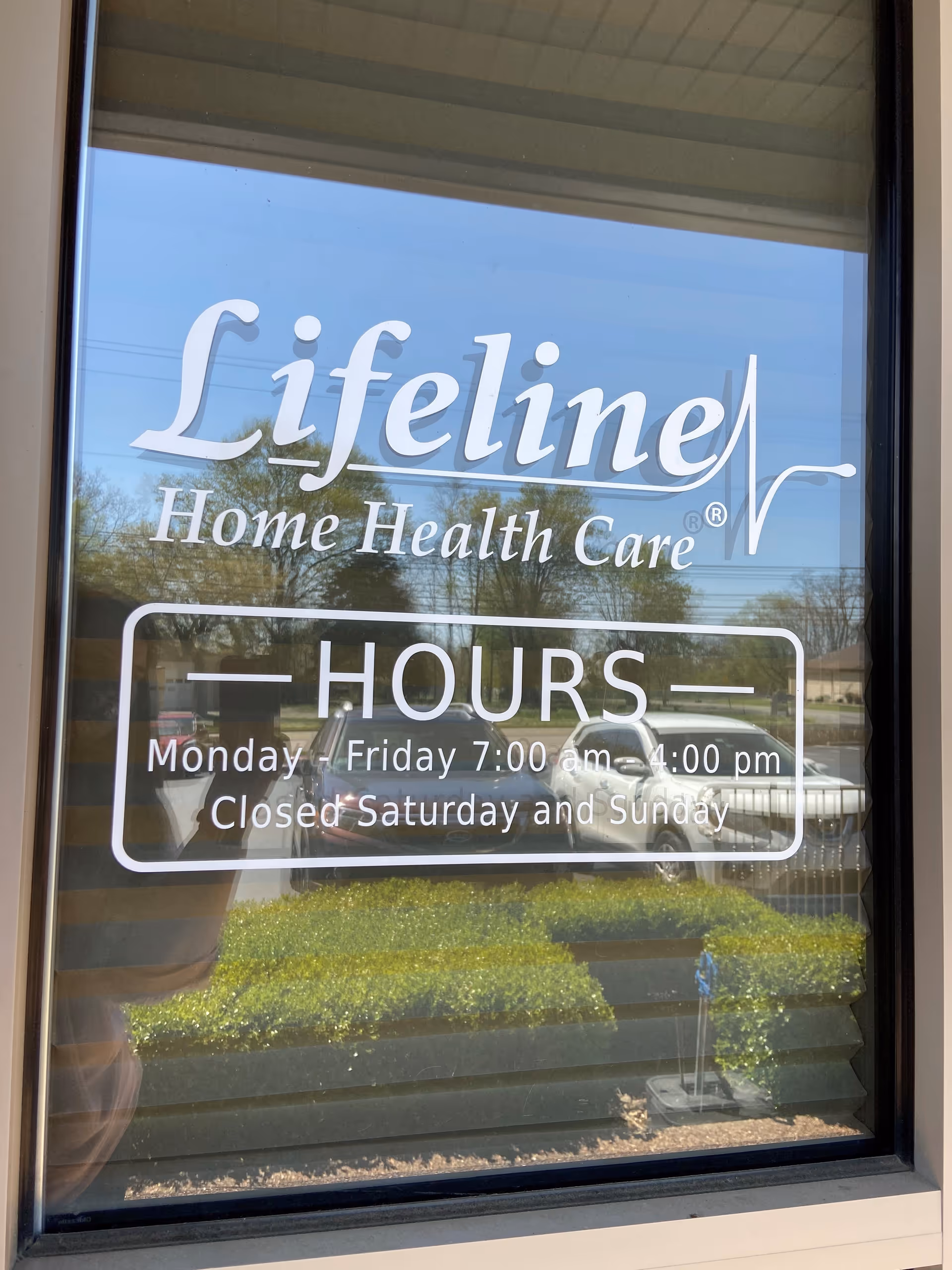 Lifeline Home Health Care sign on glass door shows hours and reflections of the parking lot.