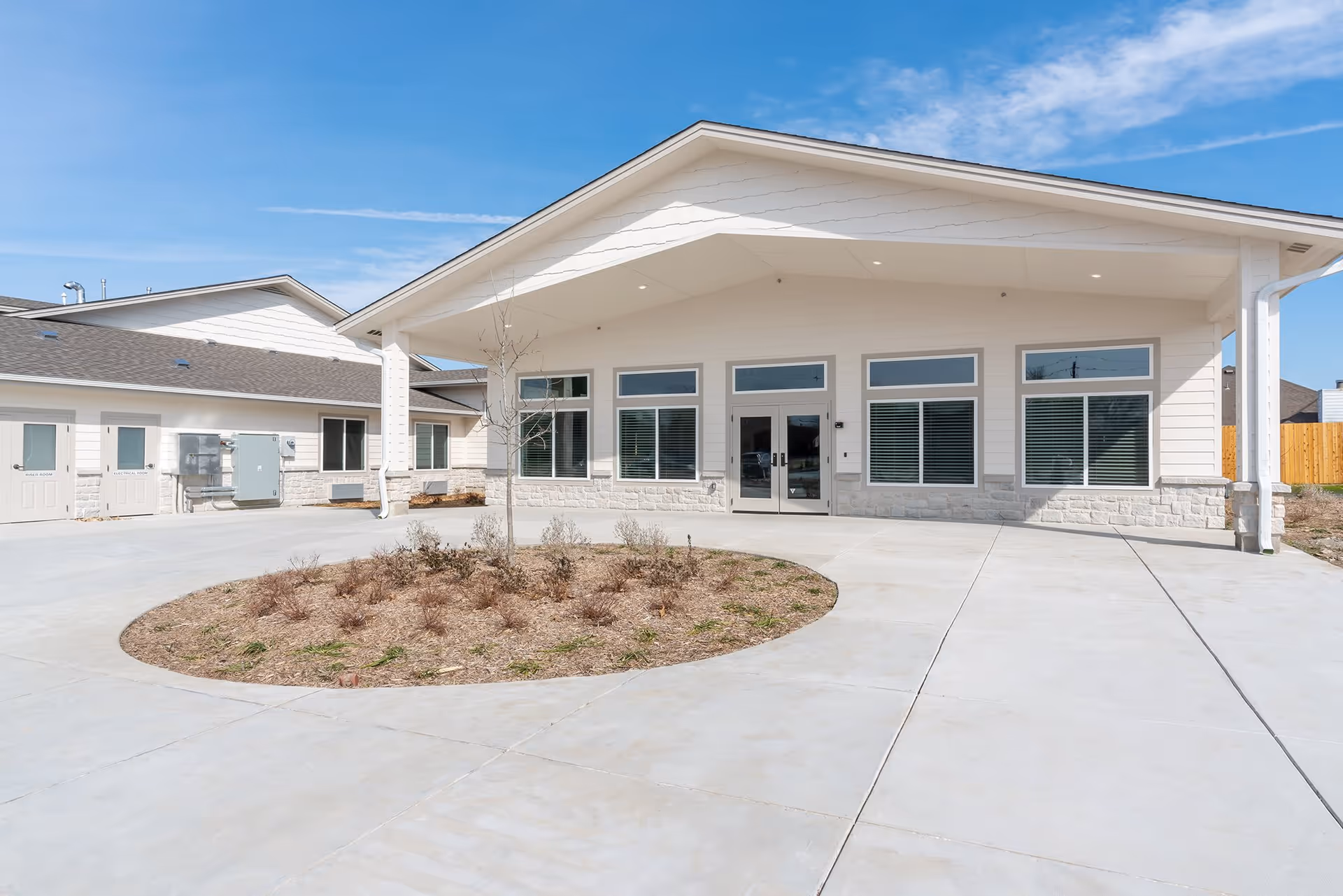 Front entrance of a single-story senior living building with a covered entry, large windows, and a circular planted island in the driveway.