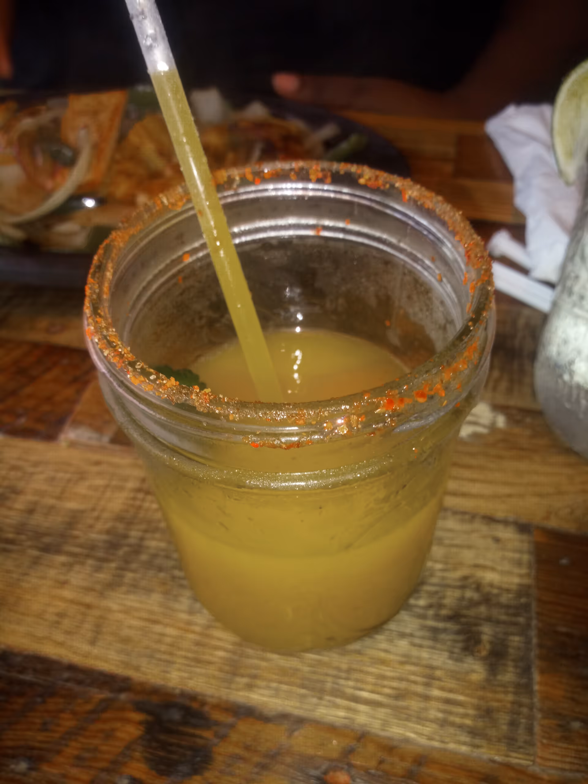Mason jar with yellow drink and straw on wooden table, background plate of food.