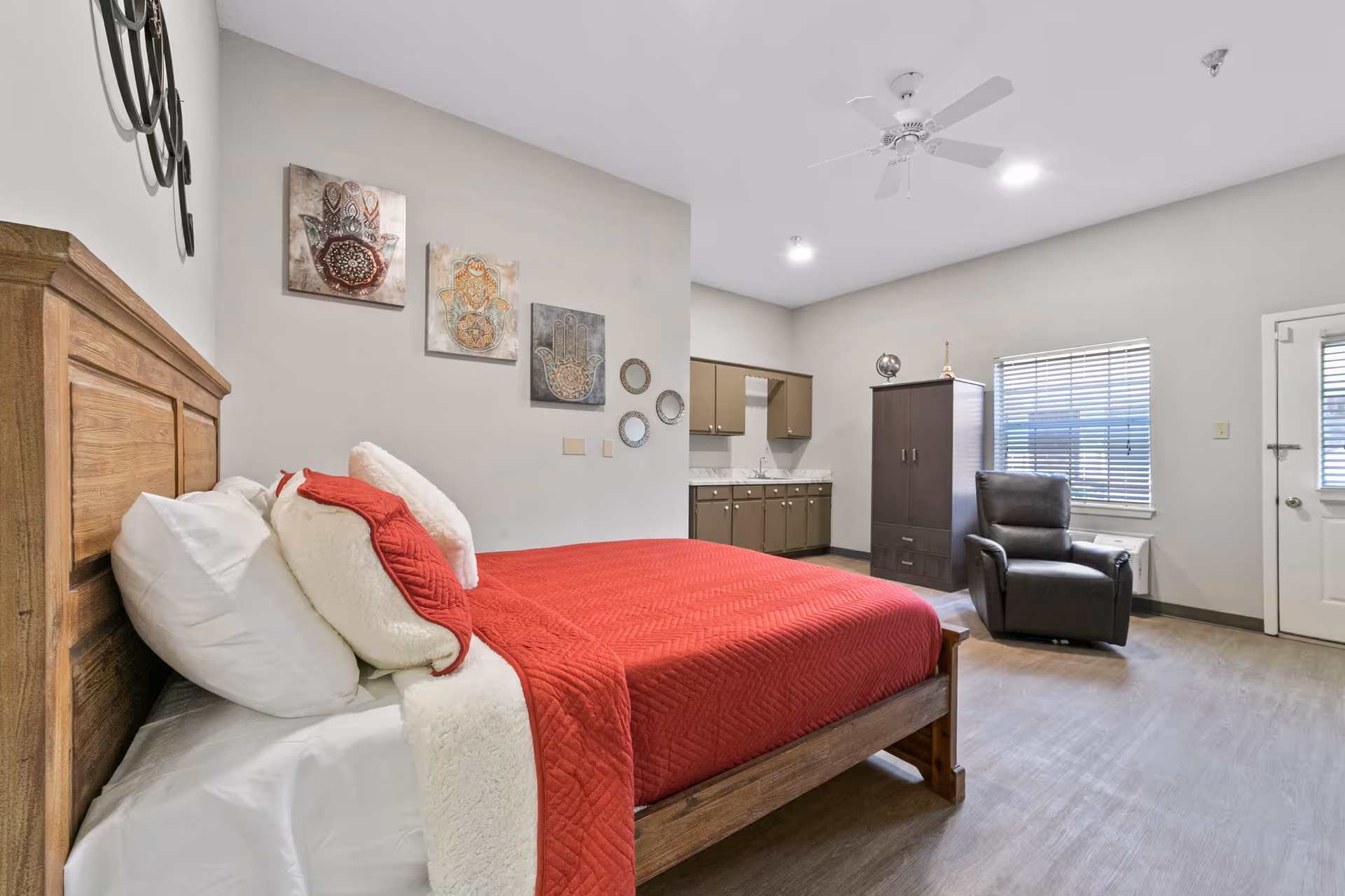 A bedroom in a senior living facility featuring a wooden bed with white pillows and a red blanket. The room has light gray walls decorated with three framed hand art pieces and three small round mirrors. There is a dark brown recliner chair near a window with blinds, a tall dark brown cabinet, and a small kitchenette area with cabinets and a sink. The floor is wood-style, and the ceiling has a white fan and recessed lighting.