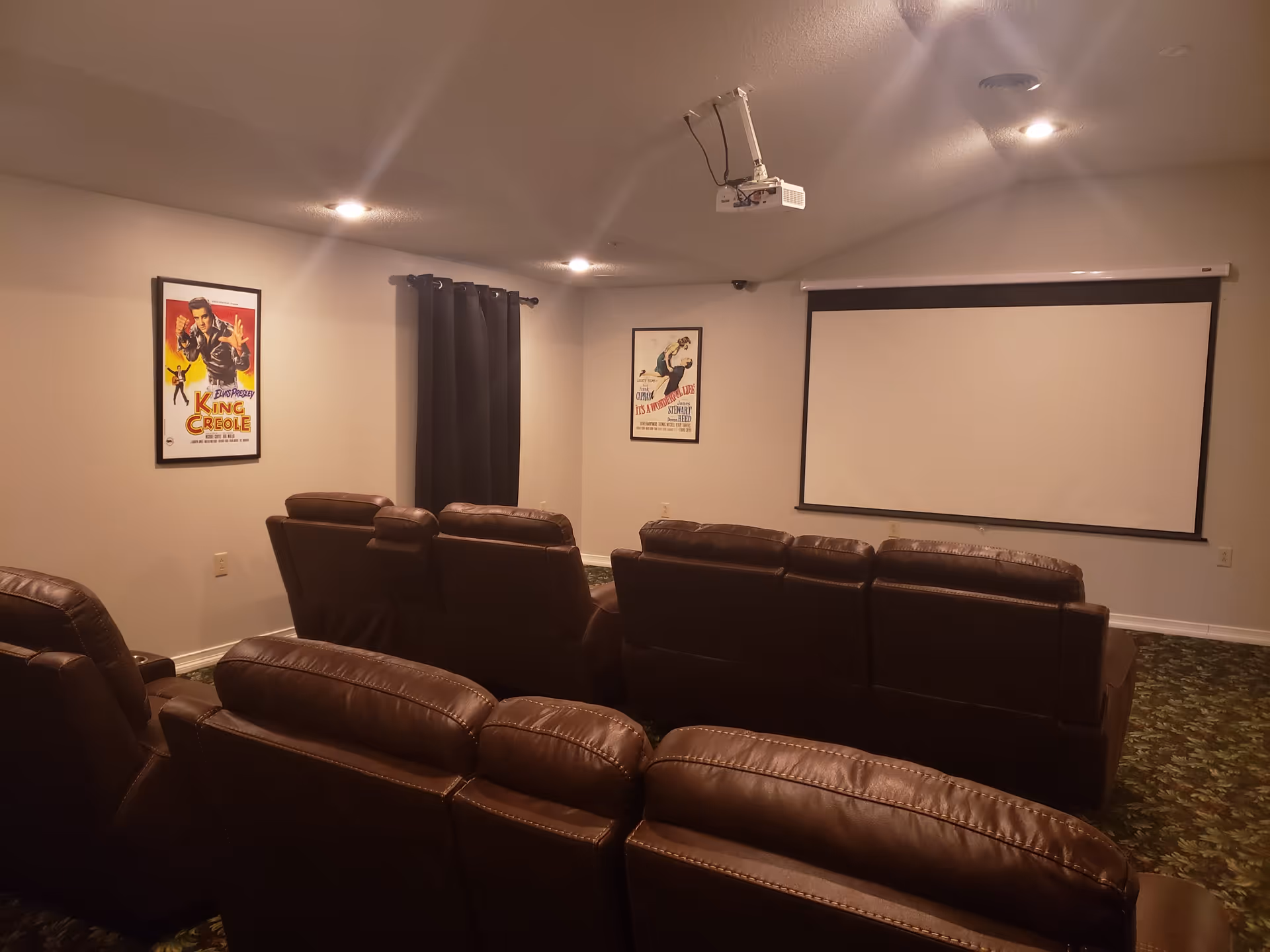 A small home theater room with two rows of brown leather recliner chairs facing a large white projector screen. The room has beige walls, a ceiling-mounted projector, two framed vintage movie posters on the walls, and a dark curtain covering a window. The floor is covered with a patterned carpet.