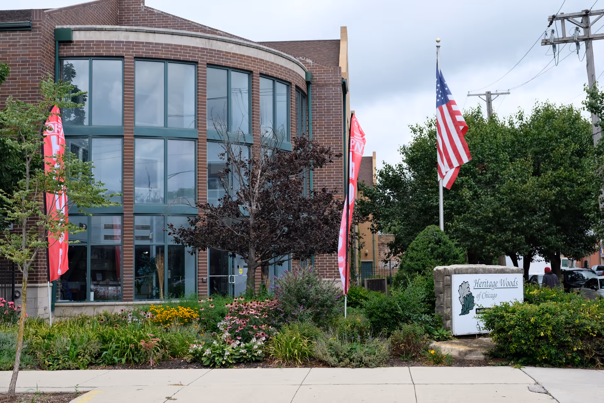 Front exterior of the Heritage Woods of Chicago building with large curved windows, landscaped garden, flagpoles, and the facility sign.