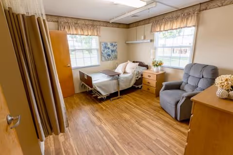 A cozy senior facility bedroom featuring a hospital bed, a recliner, and wooden dresser.