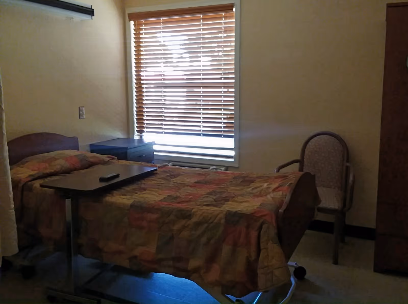A small nursing center bedroom with a single bed covered in a multicolored quilt. There is a wooden bedside table with a remote control on a movable overbed table. A window with wooden blinds is behind the bed, and a cushioned armchair is placed against the wall to the right.