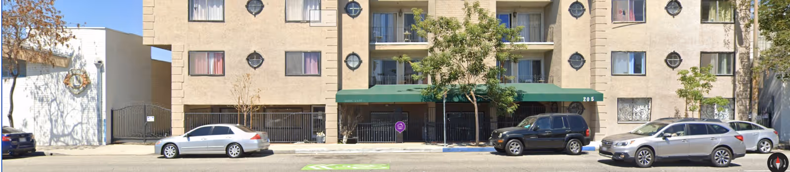 Street-level view of a multi-story apartment building with a green awning entrance, parked cars along the curb, and small trees on the sidewalk.
