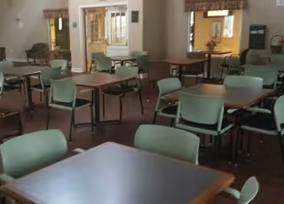 Empty dining room with multiple square tables and green chairs arranged neatly. The room has large windows with valances, and there are some chairs and a small table against the walls.