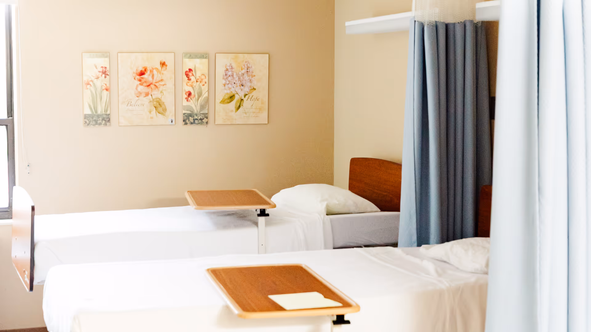 Two hospital-style beds with privacy curtains in a neutral resident room.