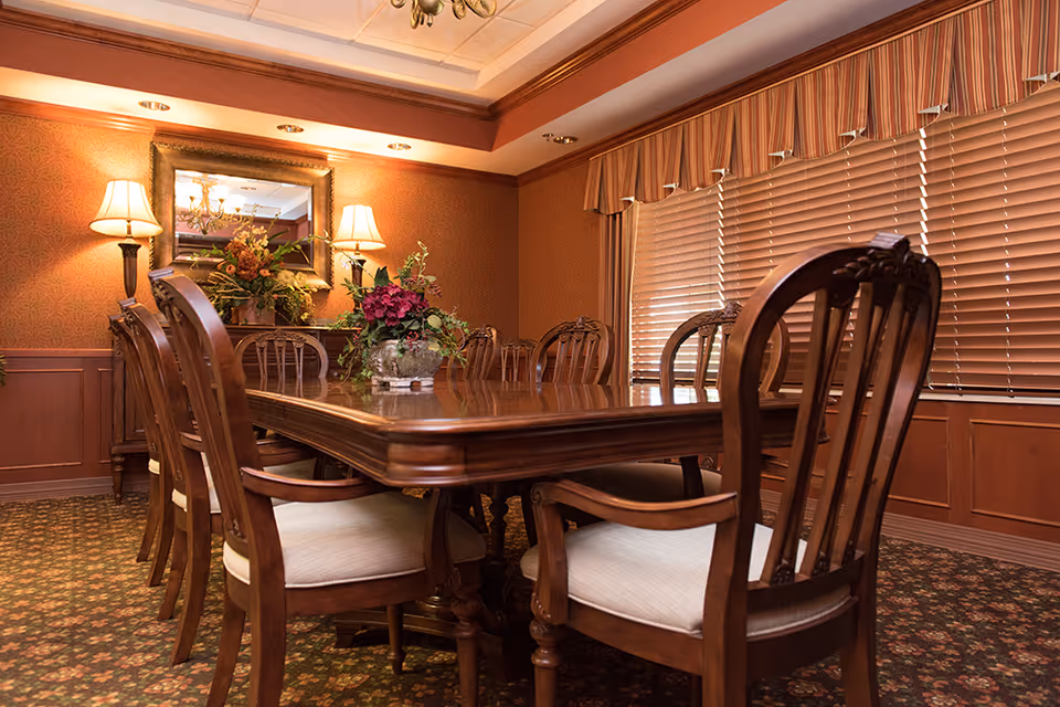 A formal dining room with a large polished wooden table surrounded by eight wooden chairs with cushioned seats. The room features patterned carpet, wood-paneled walls, a large mirror above a sideboard with floral arrangements, two wall lamps, and a window with closed blinds and a valance.