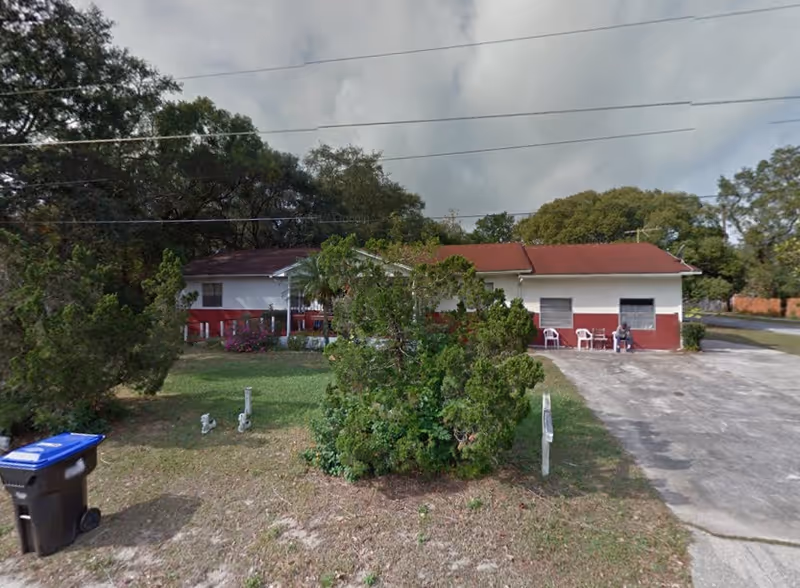 Single-story red-and-white building with a driveway, front yard trees, and porch chairs.