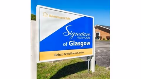 Outdoor sign for Signature HealthCARE of Glasgow Rehab & Wellness Center with a blue and white background and yellow accents, situated on grass near a parking lot and a brick building under a clear blue sky.