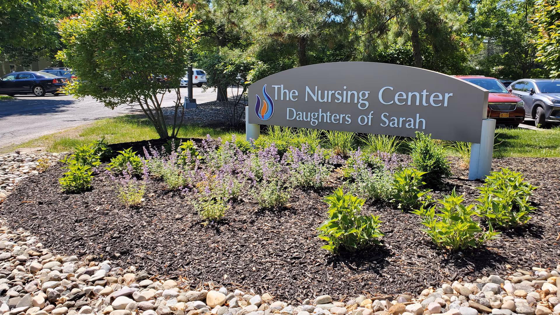 Outdoor view of a landscaped area with a sign that reads 'The Nursing Center Daughters of Sarah' surrounded by green plants and purple flowers, with parked cars and trees in the background.