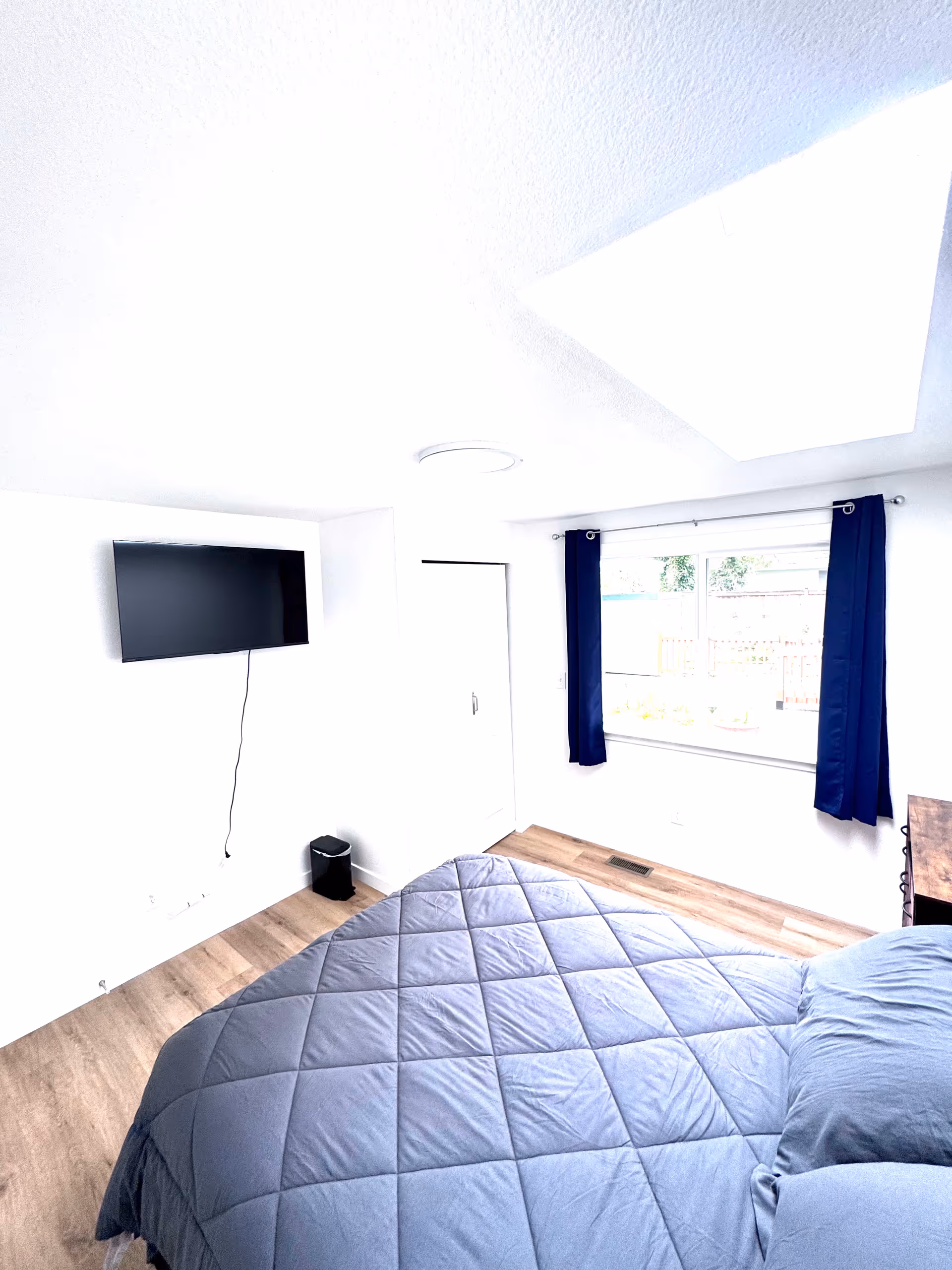A bright bedroom with a blue quilted bed, wall-mounted TV, and a window with blue curtains.