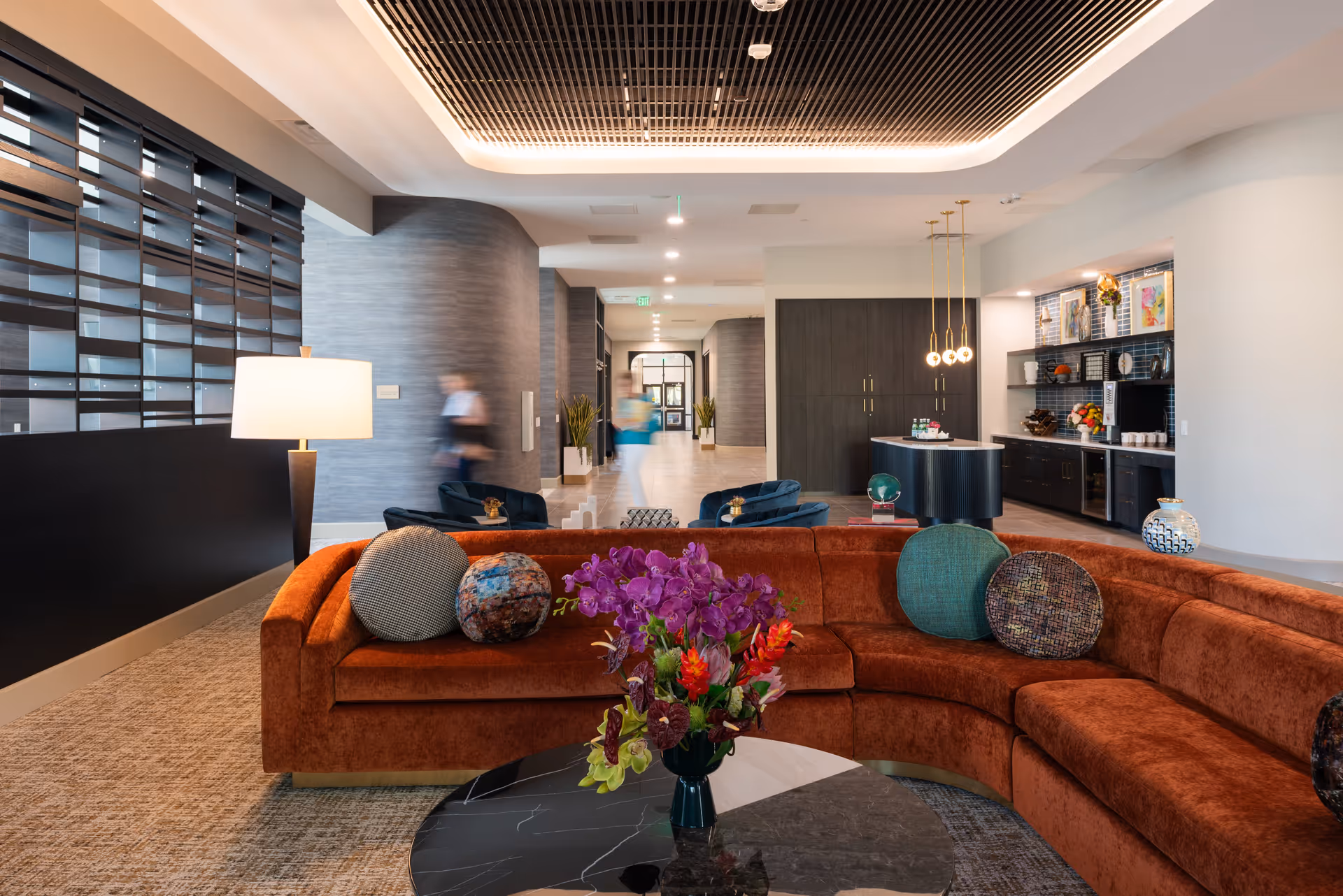 Modern senior living lounge with a curved rust-colored sectional, coffee table with flowers, and a reception/snack area in the background.
