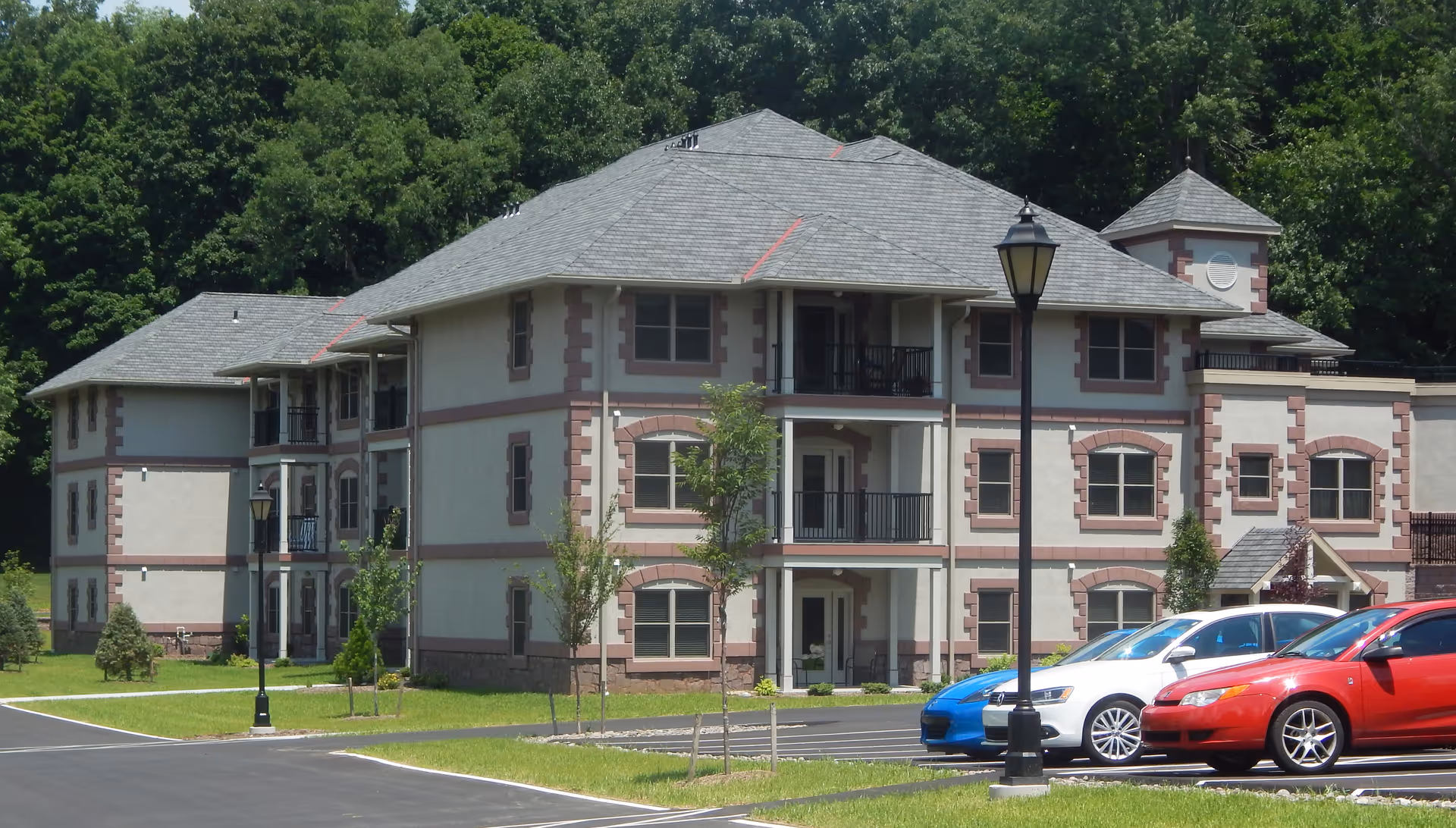 Three-story light-colored residential building with balconies and a parking lot with cars in front, surrounded by trees.
