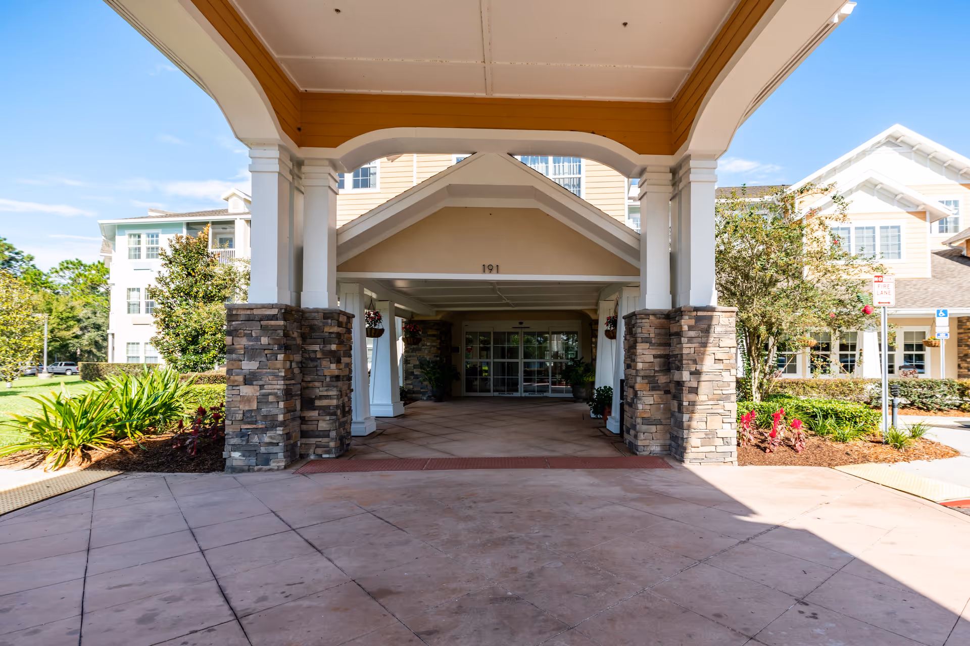 Covered entrance to a retirement living facility with stone pillars and a driveway leading to glass doors. The building is light-colored with multiple windows and surrounded by landscaped greenery and trees under a clear blue sky.
