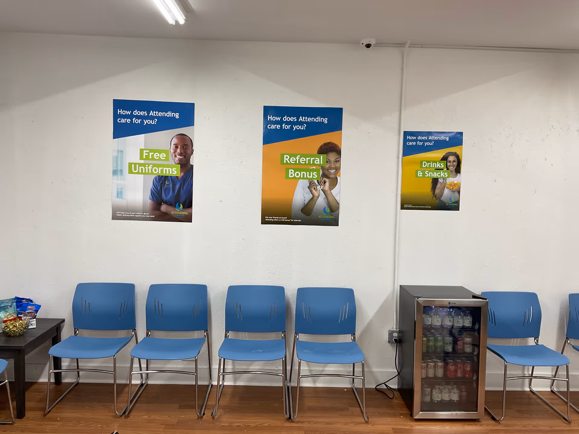 Row of blue chairs along a wall with posters and a beverage fridge in waiting area.