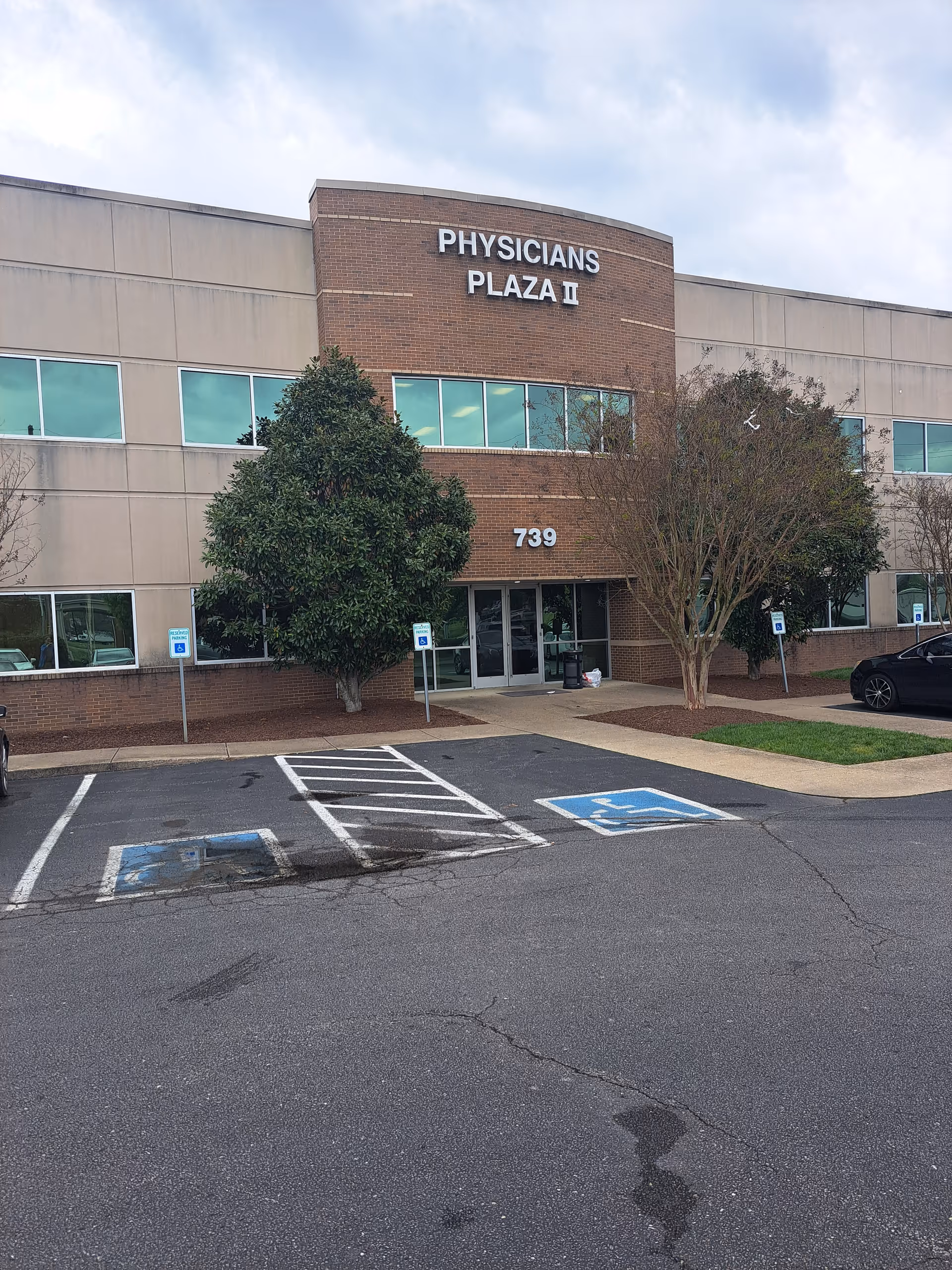 Front exterior of Physicians Plaza II medical building with parking lot and trees.