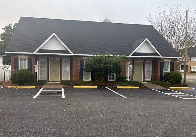 Front view of a single-story brick facility with green doors, shrubs, and parking.