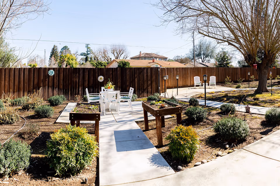 Sunny fenced courtyard with a paved walkway, raised garden beds, a white outdoor table and chairs, shrubs and trees.