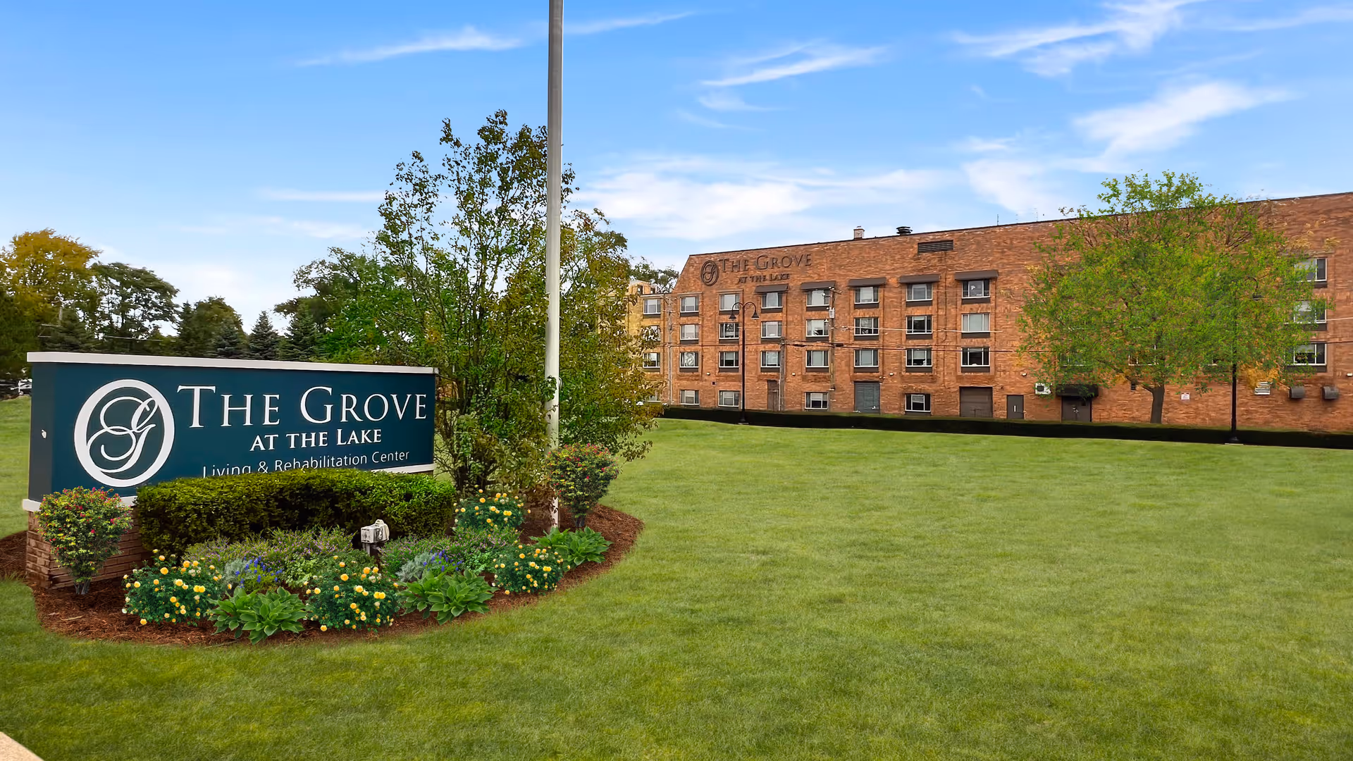 Brick senior living building with a large sign The Grove at the Lake beside a lawn.