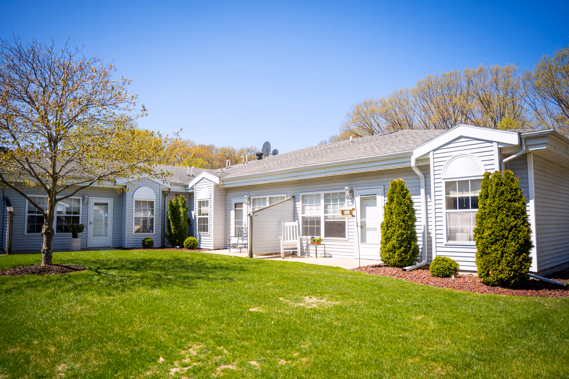 Exterior view of Woodland Terrace senior living, a single-story building with gray siding, green lawn, shrubs.