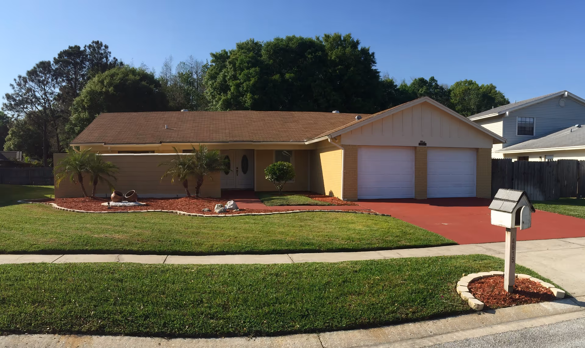 Single-story yellow brick house with a two-car garage, red driveway, front lawn, palm trees and a mailbox.