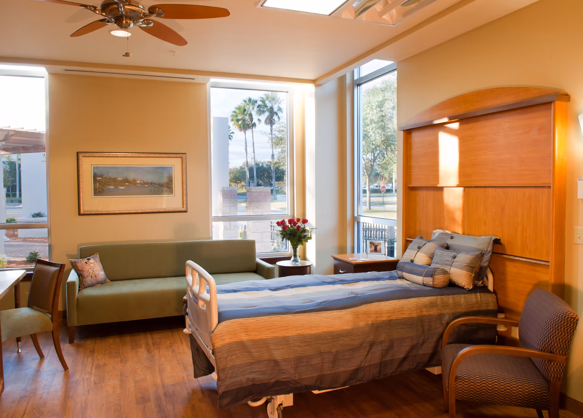Sunlit patient room with a hospital bed, green sofa, chairs, and large windows.