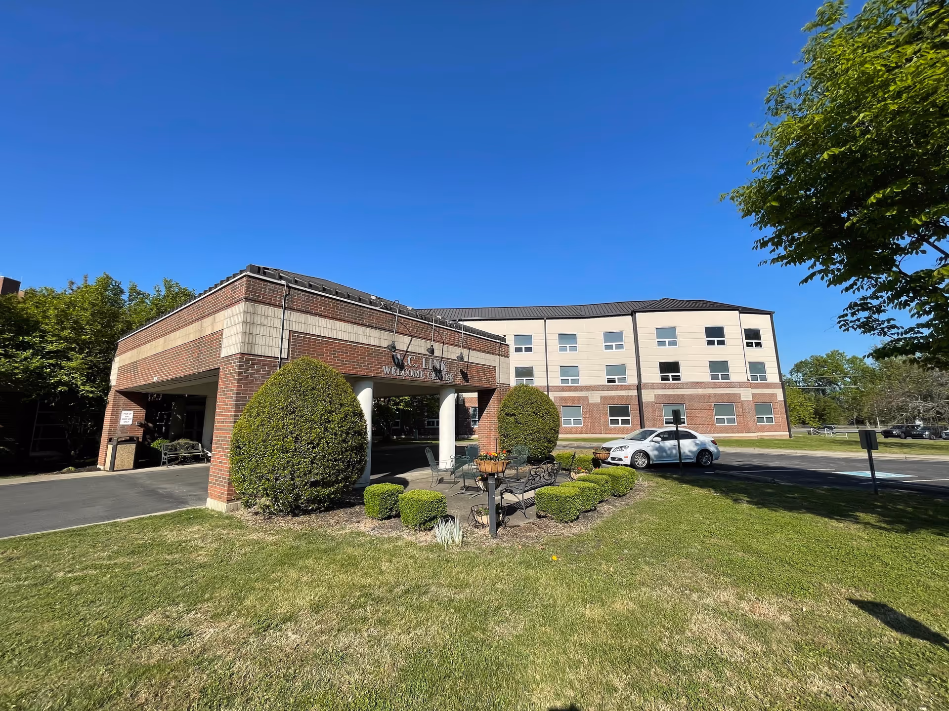 Exterior view of a multi-story brick and beige building under a clear blue sky. The building has a covered entrance with the sign 'W.C. Link Welcome Center' above it. There are neatly trimmed bushes and a small garden area with benches in front of the entrance. A white car is parked near the building, and trees surround the area.