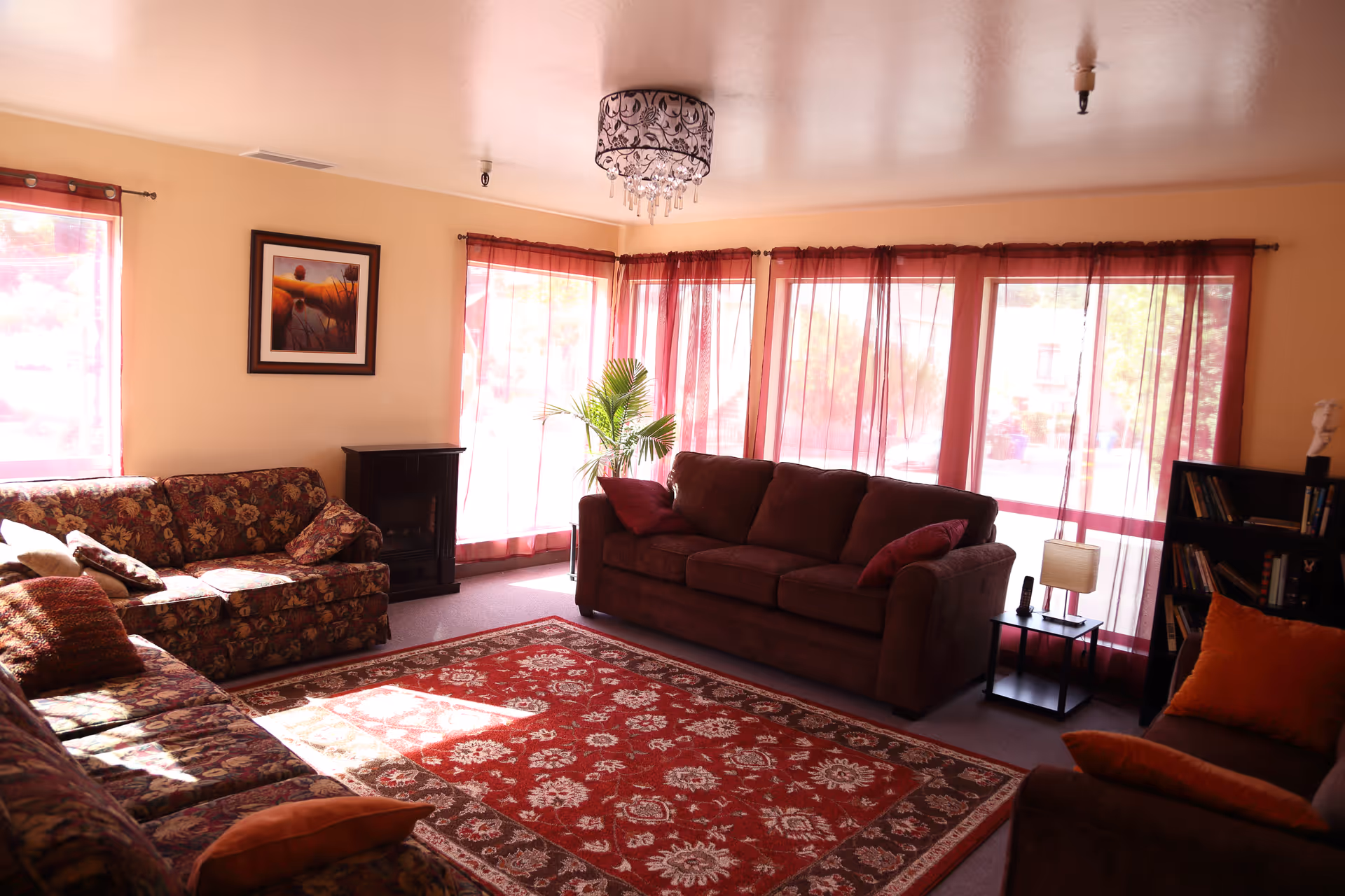 Sunlit living room with multiple sofas, a red patterned rug, sheer red curtains, and a chandelier.