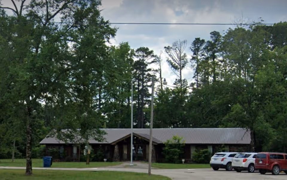 One-story, wood-clad building with a metal roof and surrounding trees in a parking lot.