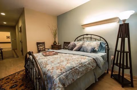 A bedroom with a metal bed, blue patterned bedding, two chairs, and a floor lamp.