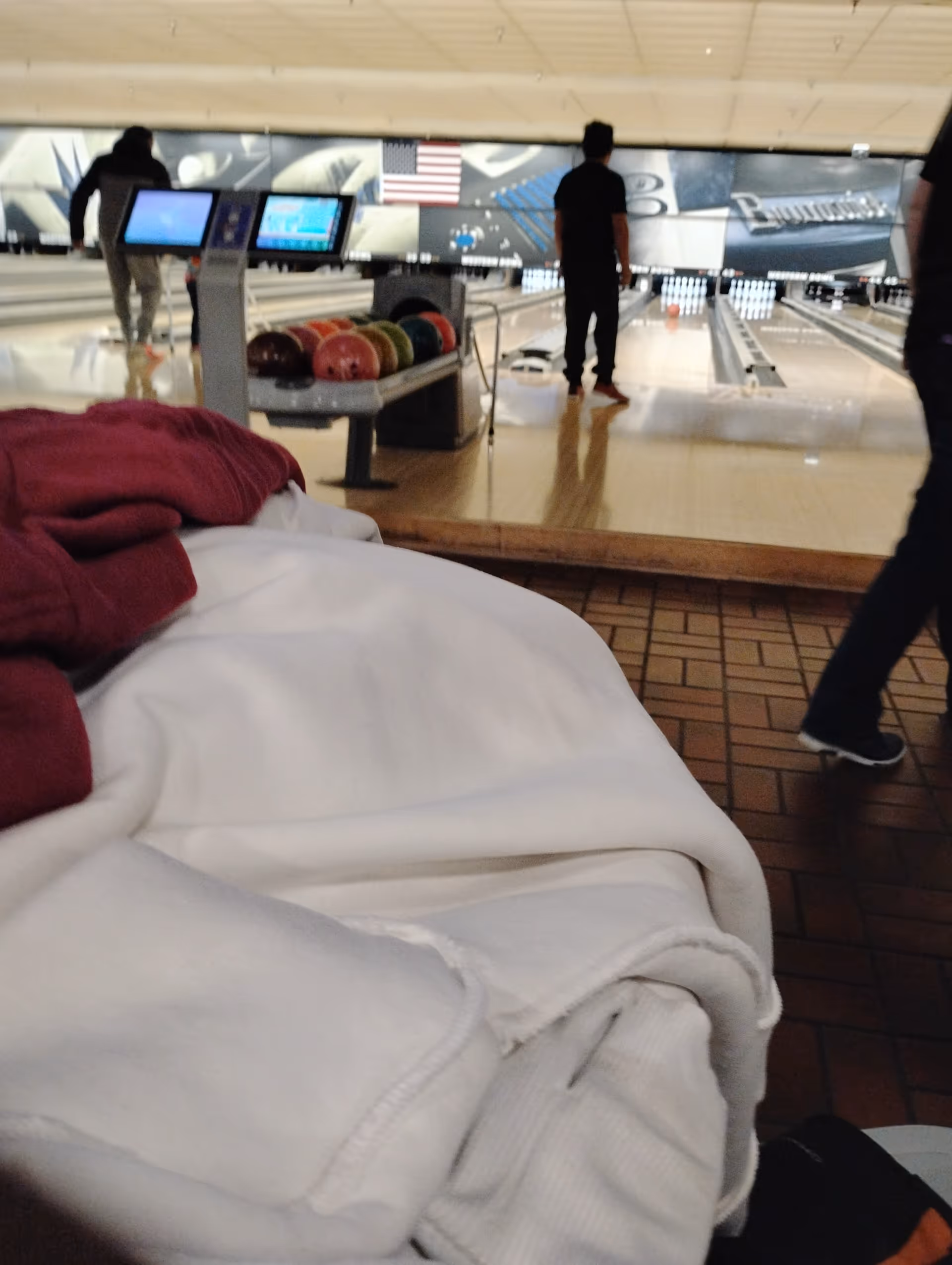 Blanket-covered bed in foreground with bowling alley and players in background.