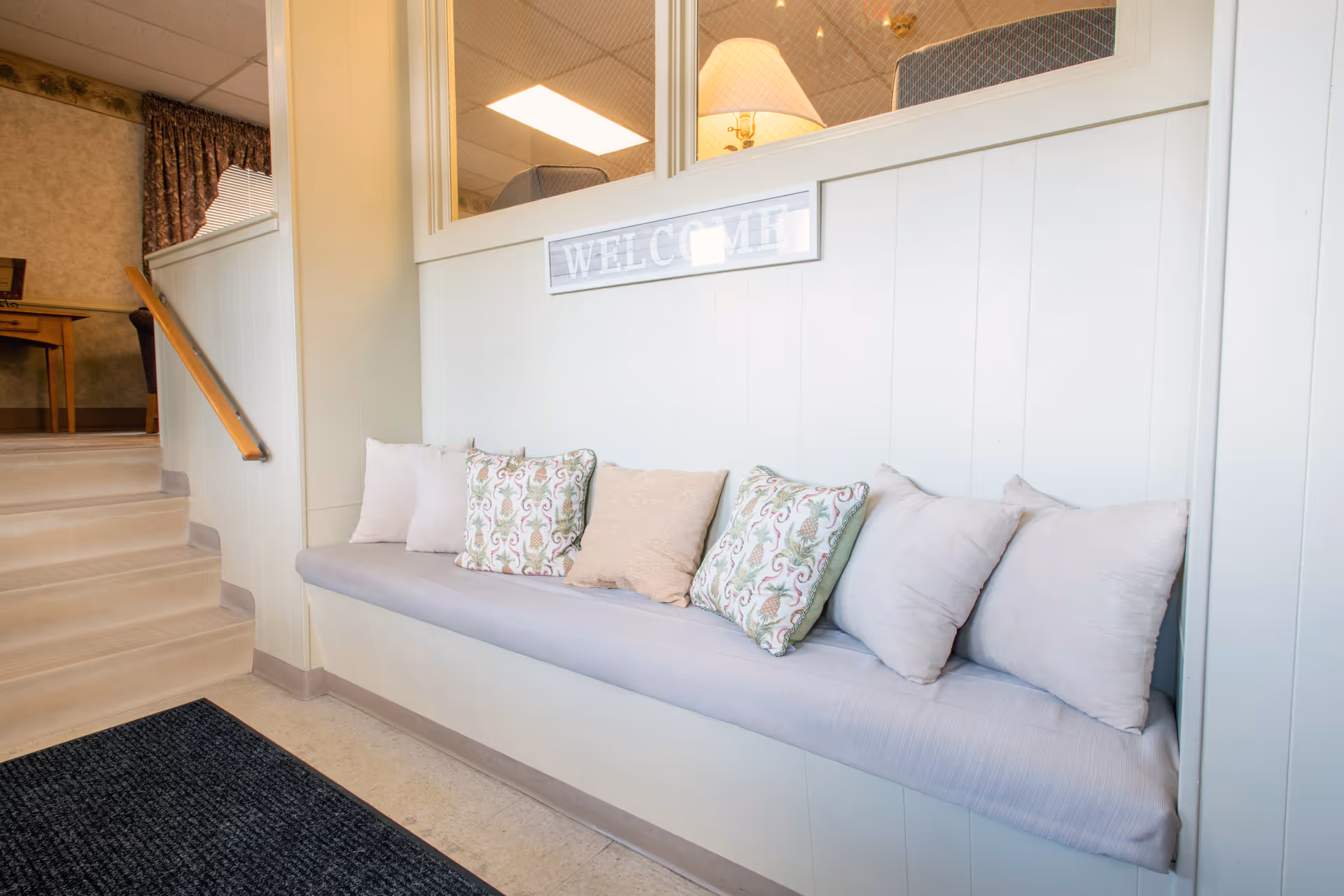 A cozy indoor seating area with a built-in bench lined with multiple cushions in light and patterned fabrics. Above the bench is a window with a 'WELCOME' sign. To the left, there is a short staircase with a wooden handrail leading to another room with a wooden table and chairs.