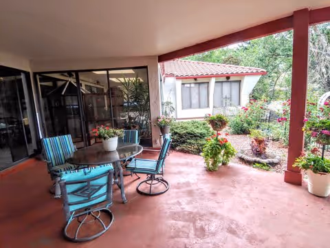 Covered outdoor patio area with a round glass table and four blue-striped cushioned chairs. The patio overlooks a garden with various plants and flowers, and a small building with a red-tiled roof is visible in the background.