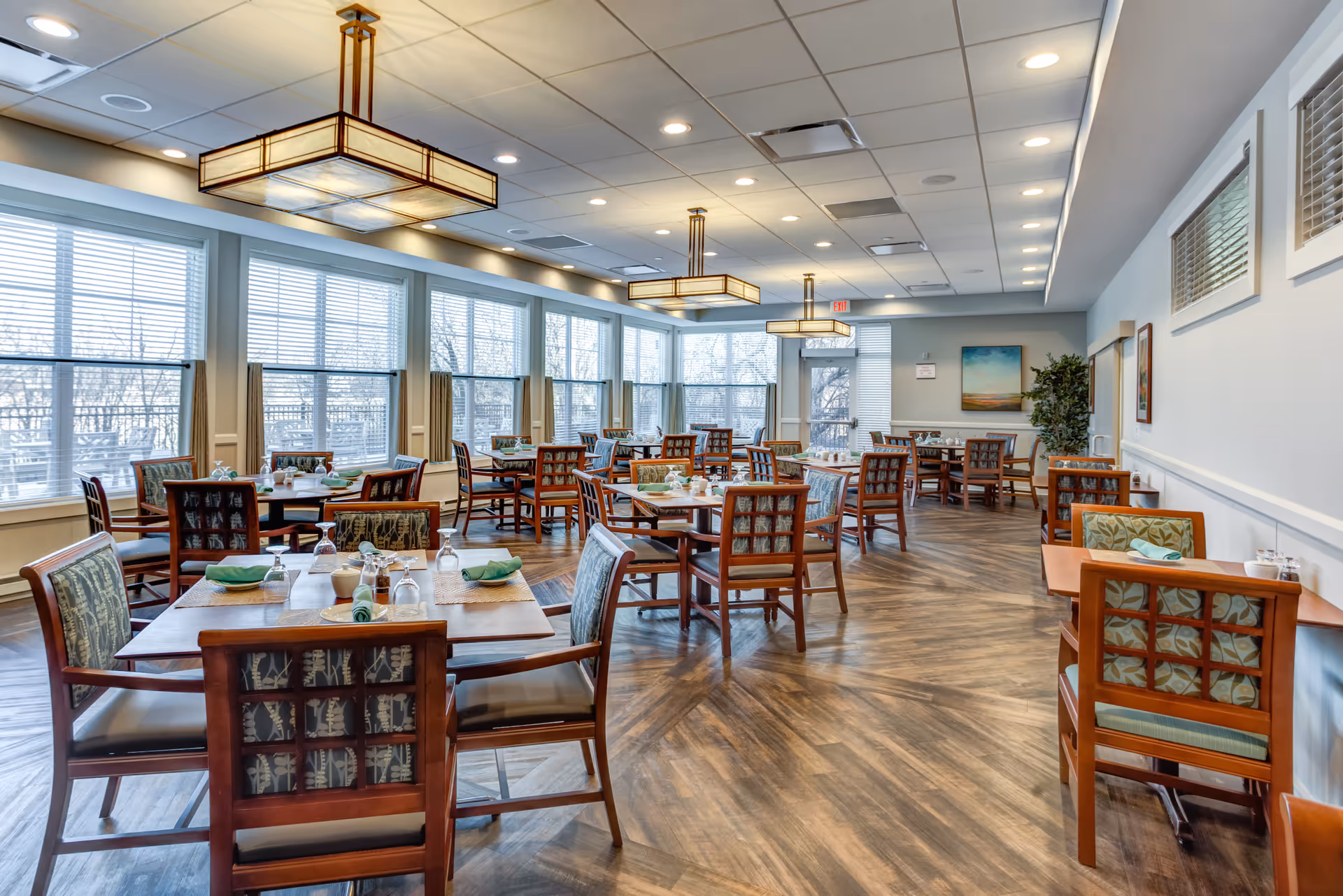 A spacious dining room with multiple wooden tables and chairs arranged neatly. Each table is set with placemats, napkins, glasses, and condiments. Large windows along one wall allow natural light to fill the room. The ceiling has modern rectangular light fixtures, and the floor features a wood-patterned design. There are paintings and a plant on the far wall.