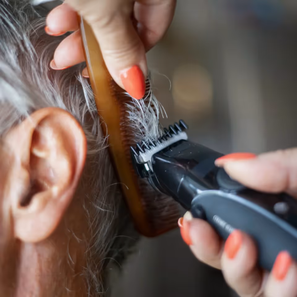 Close-up of a person trimming an elderly resident's hair with an electric clipper and a comb.