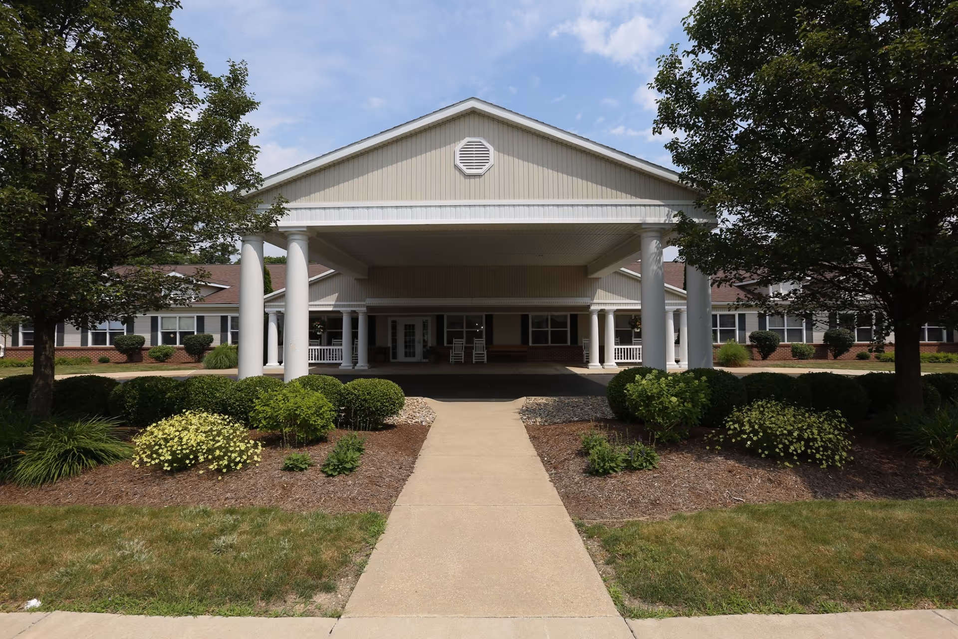 Front entrance of a one-story senior living building with a covered portico, white columns, a central walkway and landscaped shrubs.